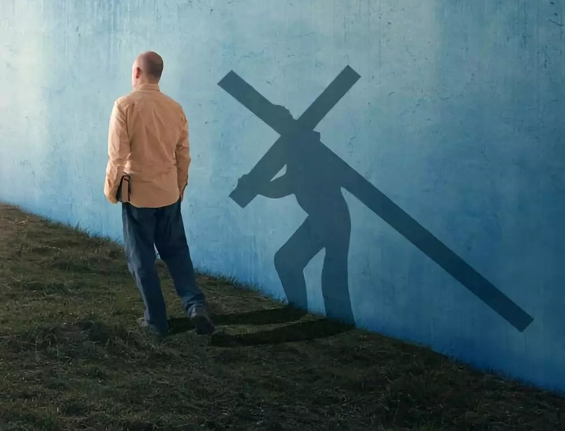 A man standing beside a wall with a Bible casts a shadow of himself carrying the cross.