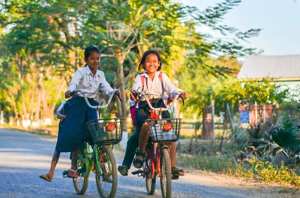 Two Cambodian students are riding bicycles in the sunset.