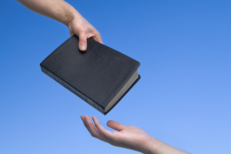 A hand is giving a Bible and another hand is receiving it.