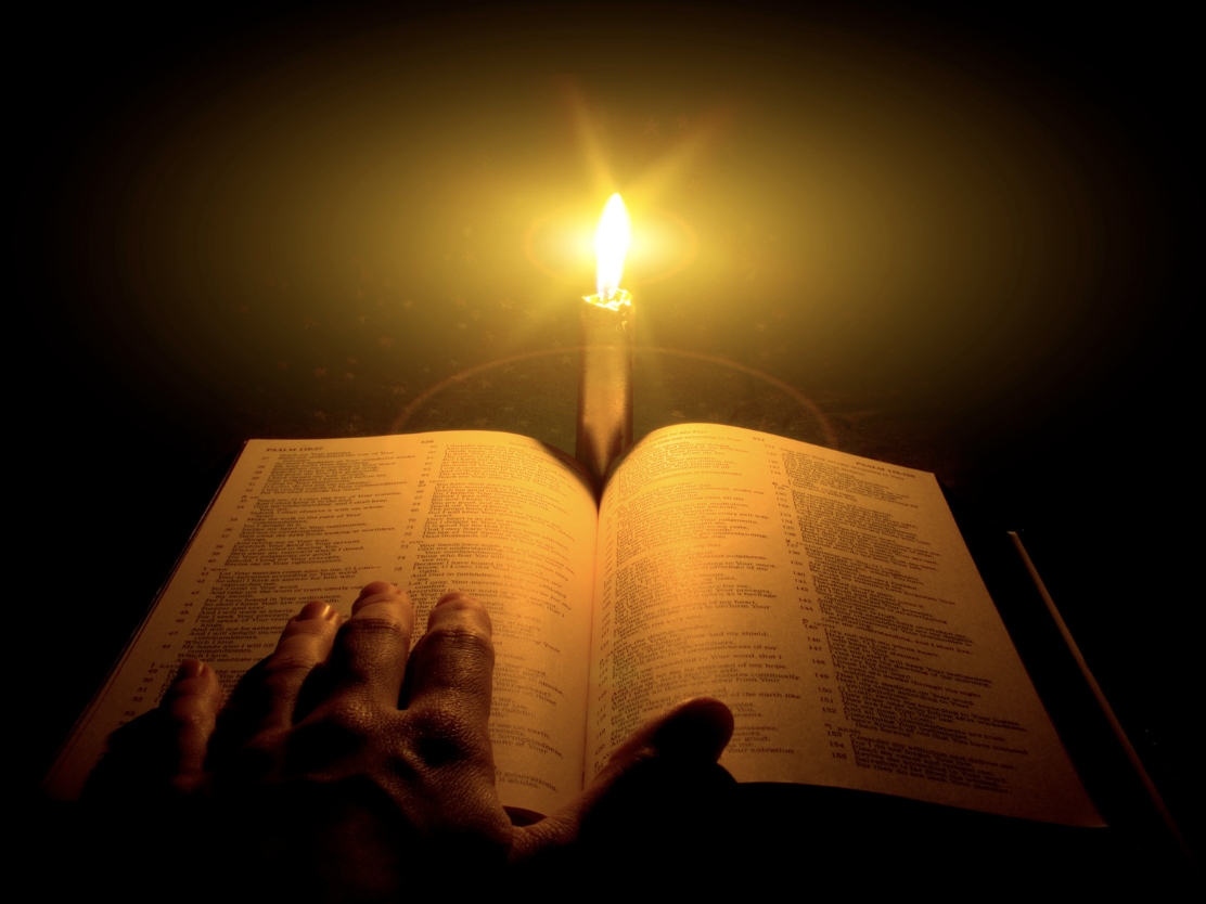 A hand is on an open Bible with a lighting candle in front. 