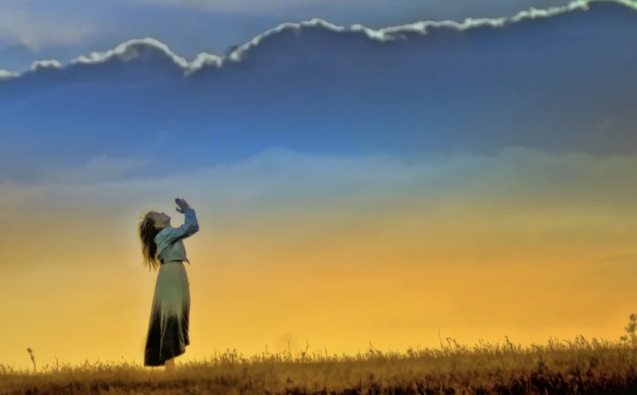 A girl prays to the sky on a hill at sunset.