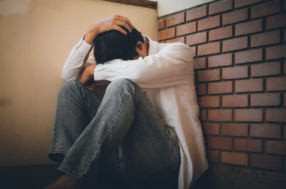 A depressed man sits with his head in hands on the floor against the wall