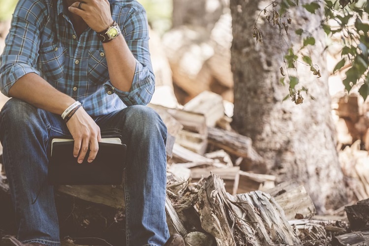 A picture of a man sitting besides a tree with a bible