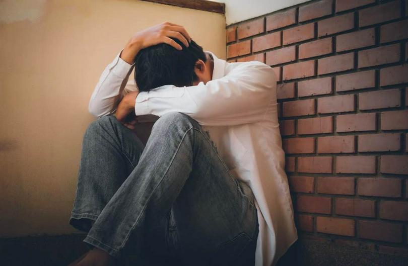 A depressed man sits with his head in hands on the floor against the wall.