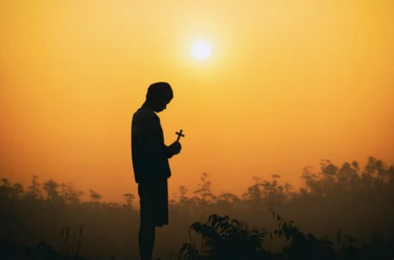 A man holds a cross and prays on the mountains at sunset. 