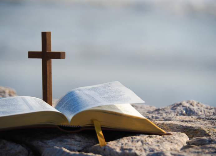 A wooden cross stands in front of an opened Bible on the rocks.