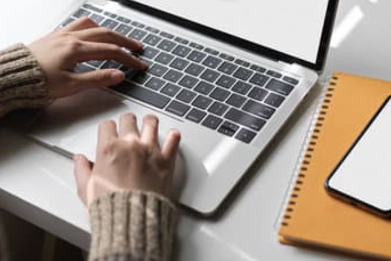 A pair of hands typed the laptop with the notebook and a mobile phone aside.